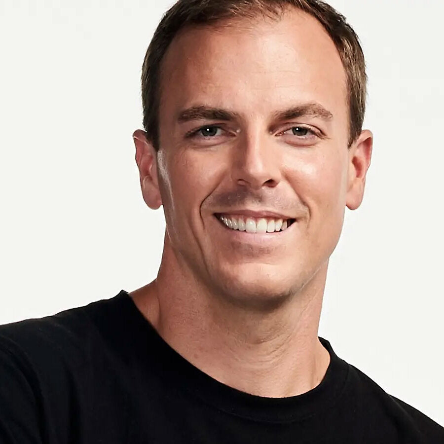 Porträt eines lächelnden Mannes mit dunklem T-Shirt vor hellem Hintergrund.