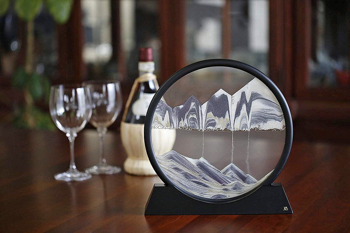 Dekoratives, rundes Glas mit Berg- und Wasserreflexion, auf Holzplatte, im Hintergrund Wein- und Spirituosenflasche, Gläser.