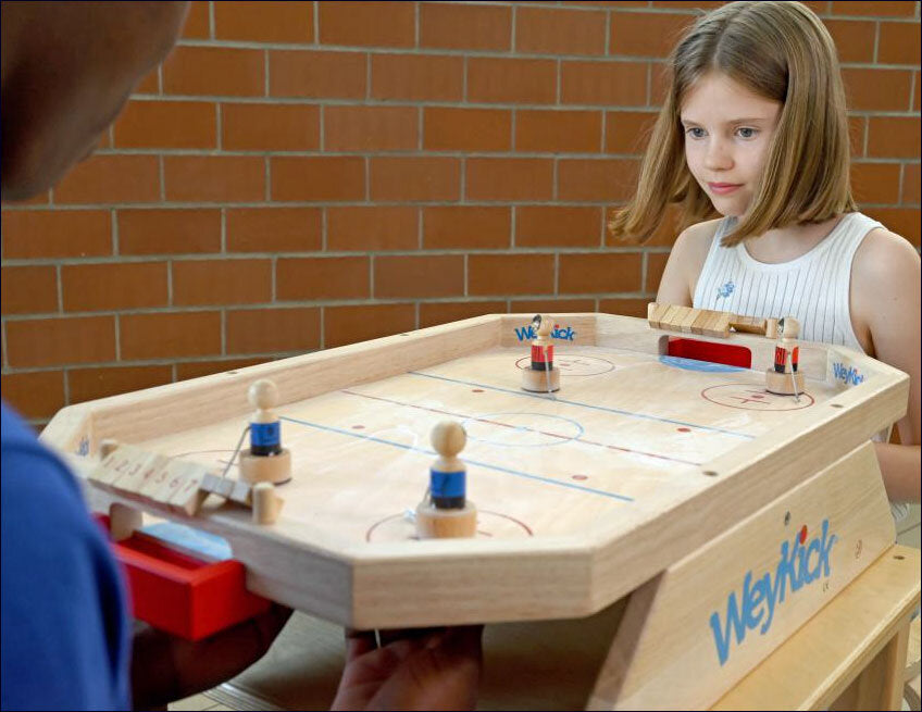 Junge und Erwachsener spielen Tischhockey auf einem Holzspielbrett vor einer Ziegelwand.