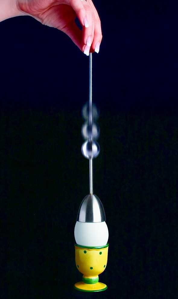 Hand hält Pendel, das über einem Eierkopf-Objekt schwingt, gegen schwarzen Hintergrund.