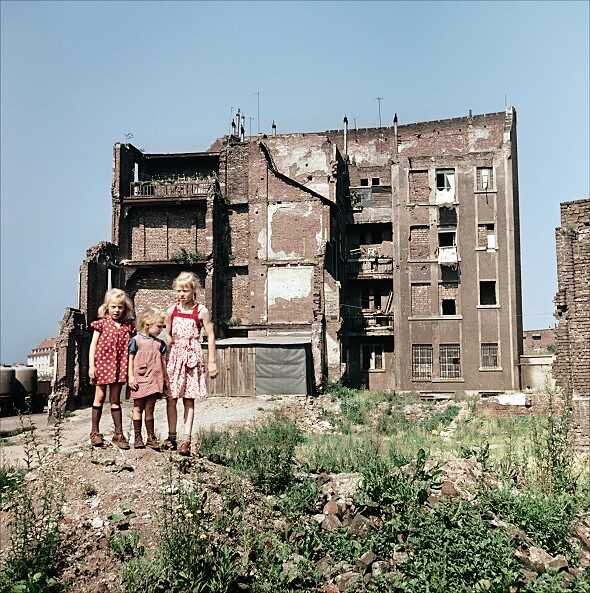 Drei Kinder stehen vor einem zerstörten, bröckelnden Mehrfamilienhaus mit beschädigten Fenstern und Fassaden.