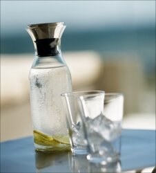 Glasvase mit Wasser und Zitronenscheiben, daneben zwei leere Gläser auf einem Tisch, im Hintergrund unscharfer Ausblick.