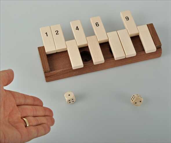 Holzblock mit nummerierten Klötzen, zwei Würfel (eine mit sechs und eine mit fünf Seiten) und eine Hand mit Ring.