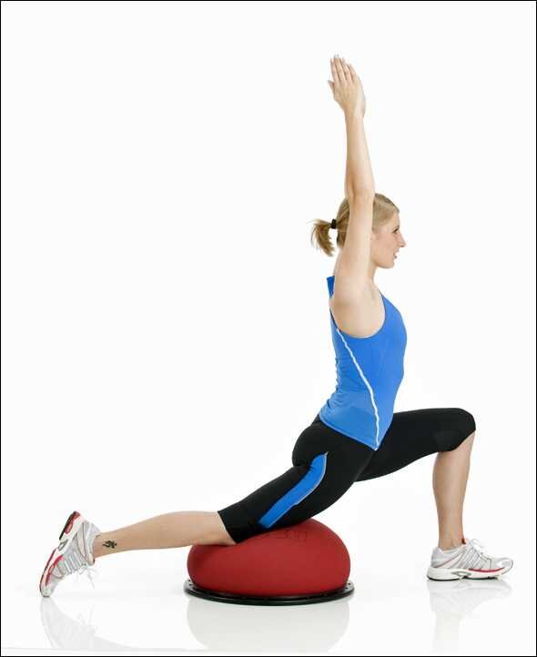 Frau trainiert auf einem Balanceball, in einer tiefen Ausfallschritt-Position mit erhobenen Armen.