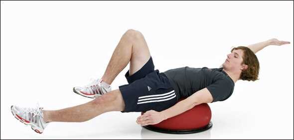Mann liegt seitlich auf einem Balance-Board, mit einem roten Ball unter dem Rücken, Arme gestreckt nach oben.