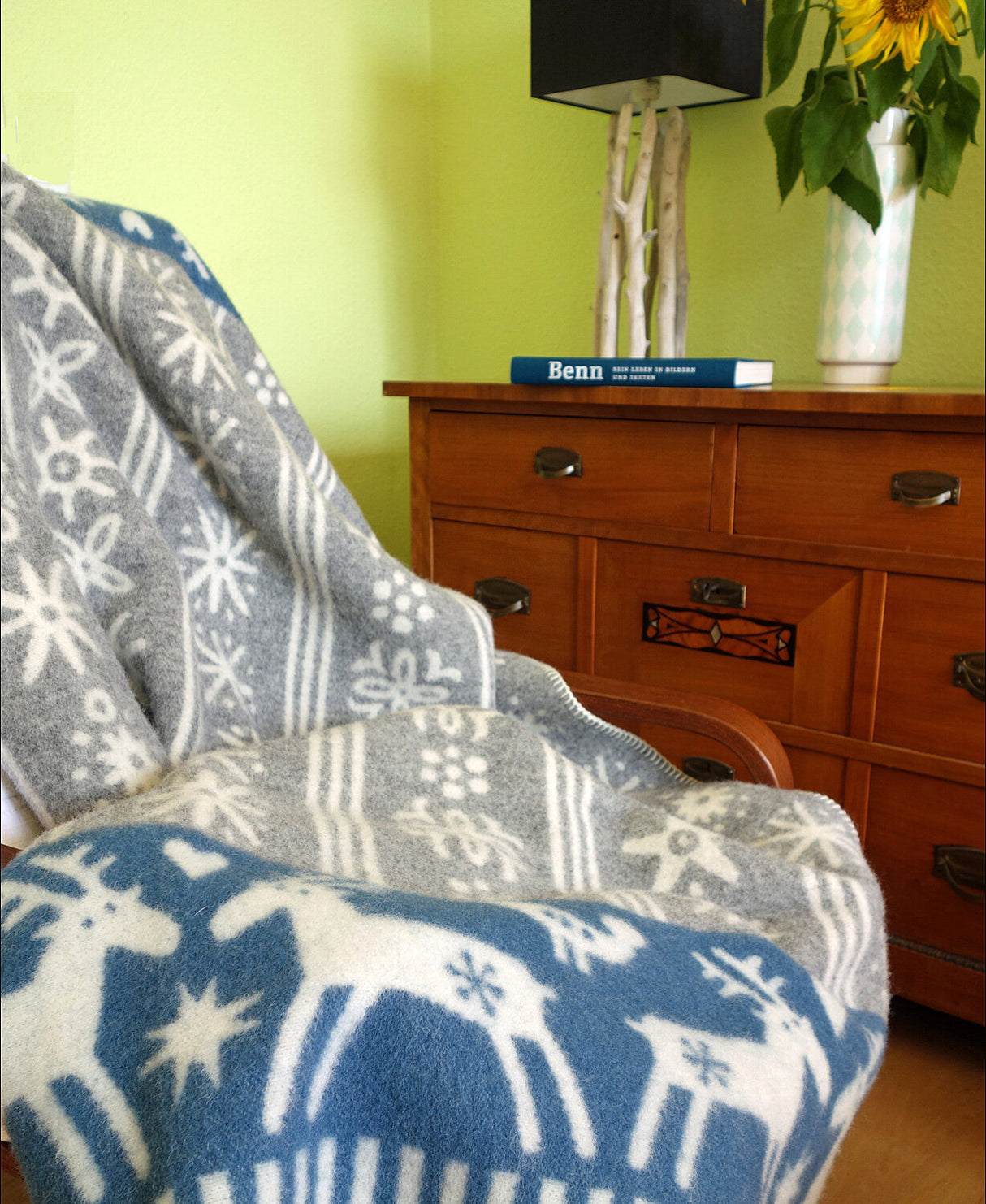 Blick auf eine Couch mit grauem, weihnachtlich gemustertem Decke, Holzkommode mit Buch, Vase mit Sonnenblume und Lampen