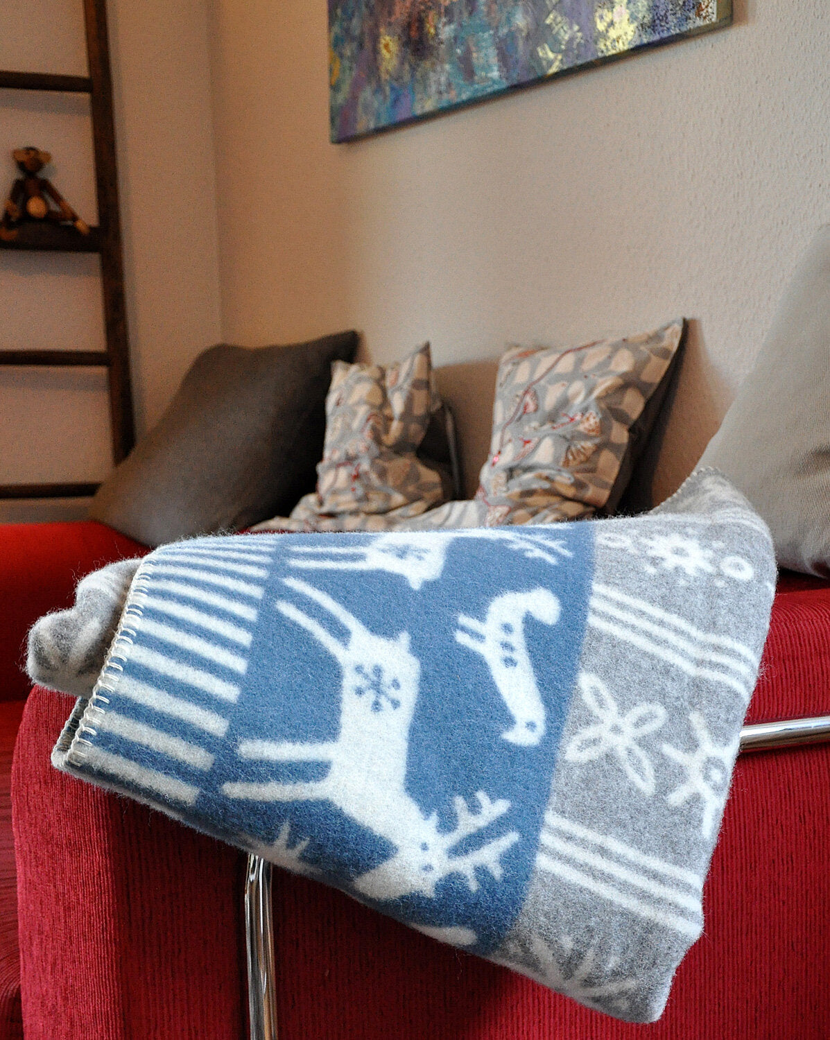 Wollenergiematte mit Rentier- und Schneeflocken-Muster liegt auf rotem Sofa, im Hintergrund Kissen und Wandbild.
