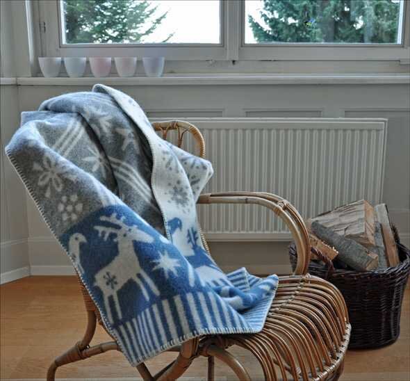 Wickerstuhl mit grauem und blauen Weihnachtsdecke, vor einem Fenster mit weißen Blumentöpfen, daneben ein Korb mit Holzscheiten.
