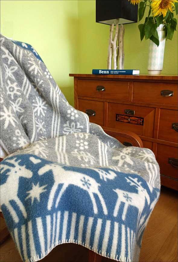 Sessel mit grauem und blauen Decke mit Schneeflocken- und Rentier-Muster, Holzkommode mit Buch und Vase mit Sonnenblumen.