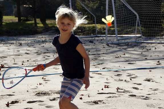 Ein lachendes Mädchen mit langen Haaren, das im Sand auf einem Tennisplatz mit einem Schläger spielt.