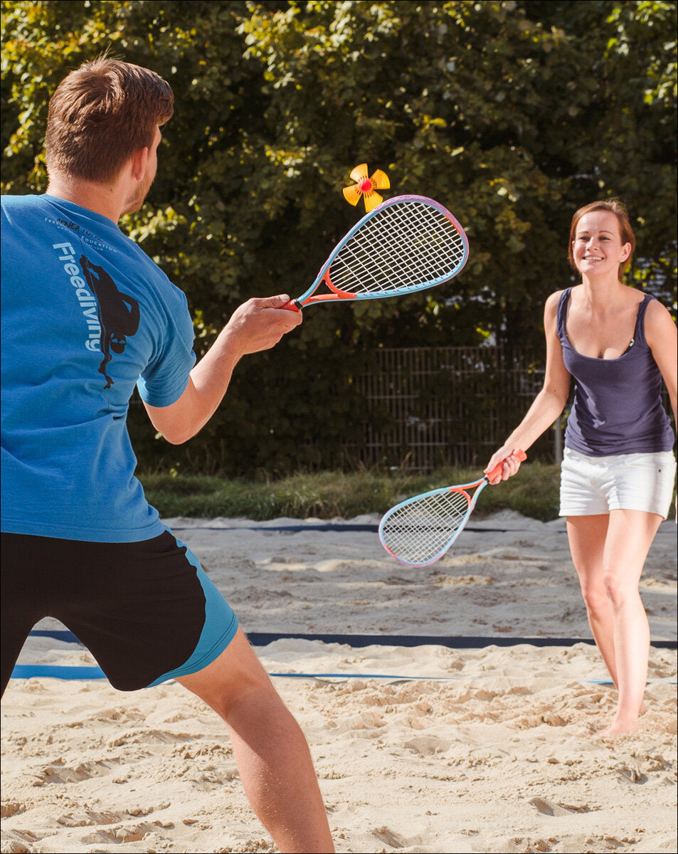 Zwei Personen spielen Beachtennis auf Sand, mit einem Windrad als Ball.