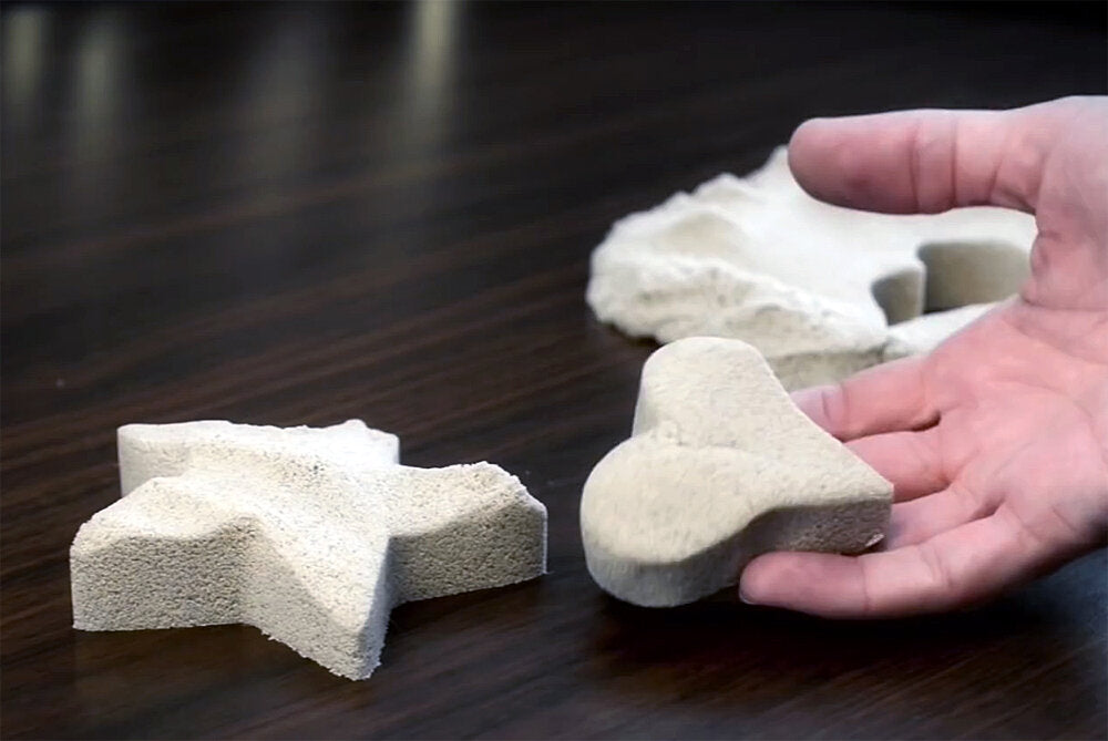 Hand hält ein unvollständiges Puzzlestück aus hellem Stein, weitere Puzzlestücke liegen auf dunklem Tisch.