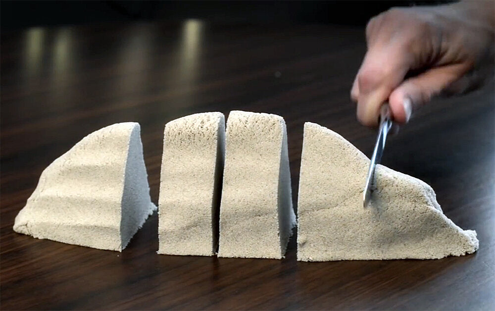 Vier große, weiße, dreieckige Objekte auf einem dunklen Tisch, eine Hand schneidet eines mit einem Messer.