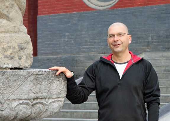 Mann mit Brille, schwarzer Jacke, weißem Hemd, steht vor einer Treppe und berührt eine große Steinfigur.