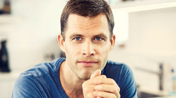 Nahaufnahme eines Mannes mit kurzen dunklen Haaren, blauen Augen und blauer Kleidung, der direkt in die Kamera schaut.