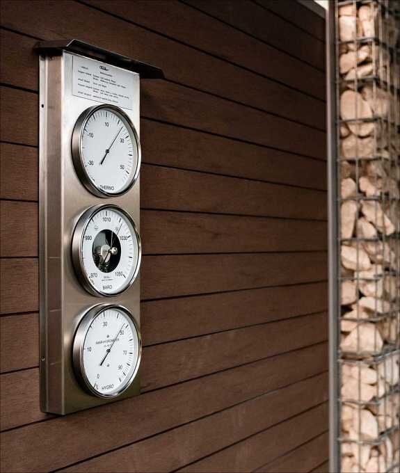 Wetterstation mit drei runden Messinstrumenten an einer Holzfassade, daneben ein Steinkunstwerk.