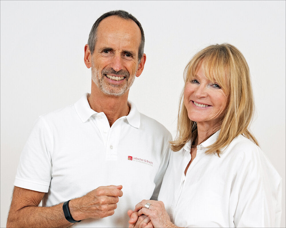 Ein Mann und eine Frau, beide in weißen Hemden, stehen vor weißem Hintergrund und lächeln.