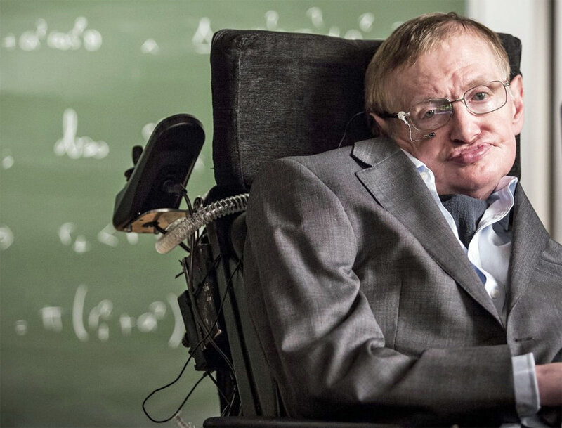 Mann im Rollstuhl vor einer Tafel mit mathematischen Formeln, trägt Anzug und Brille, wirkt nachdenklich.