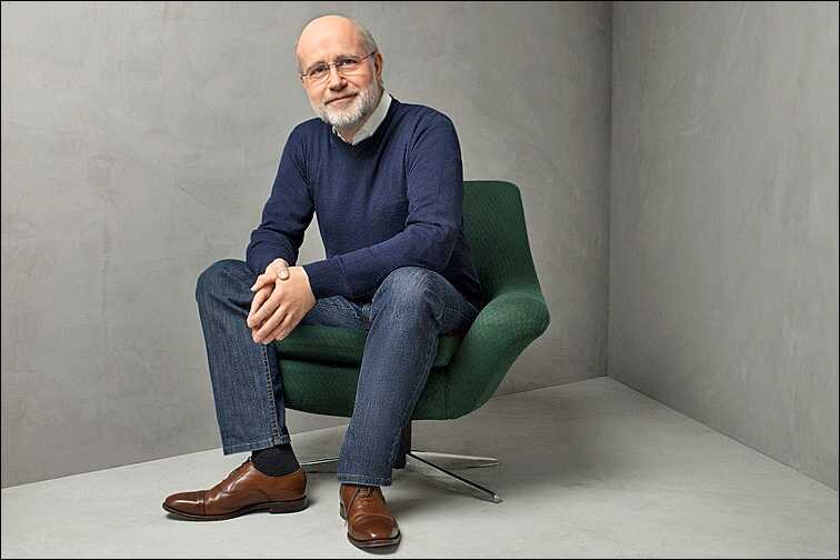 Älterer Mann mit Brille, grauem Bart, in dunklem Pullover, Jeans und braunen Schuhen sitzt auf grünem Sessel in grauem Raum.
