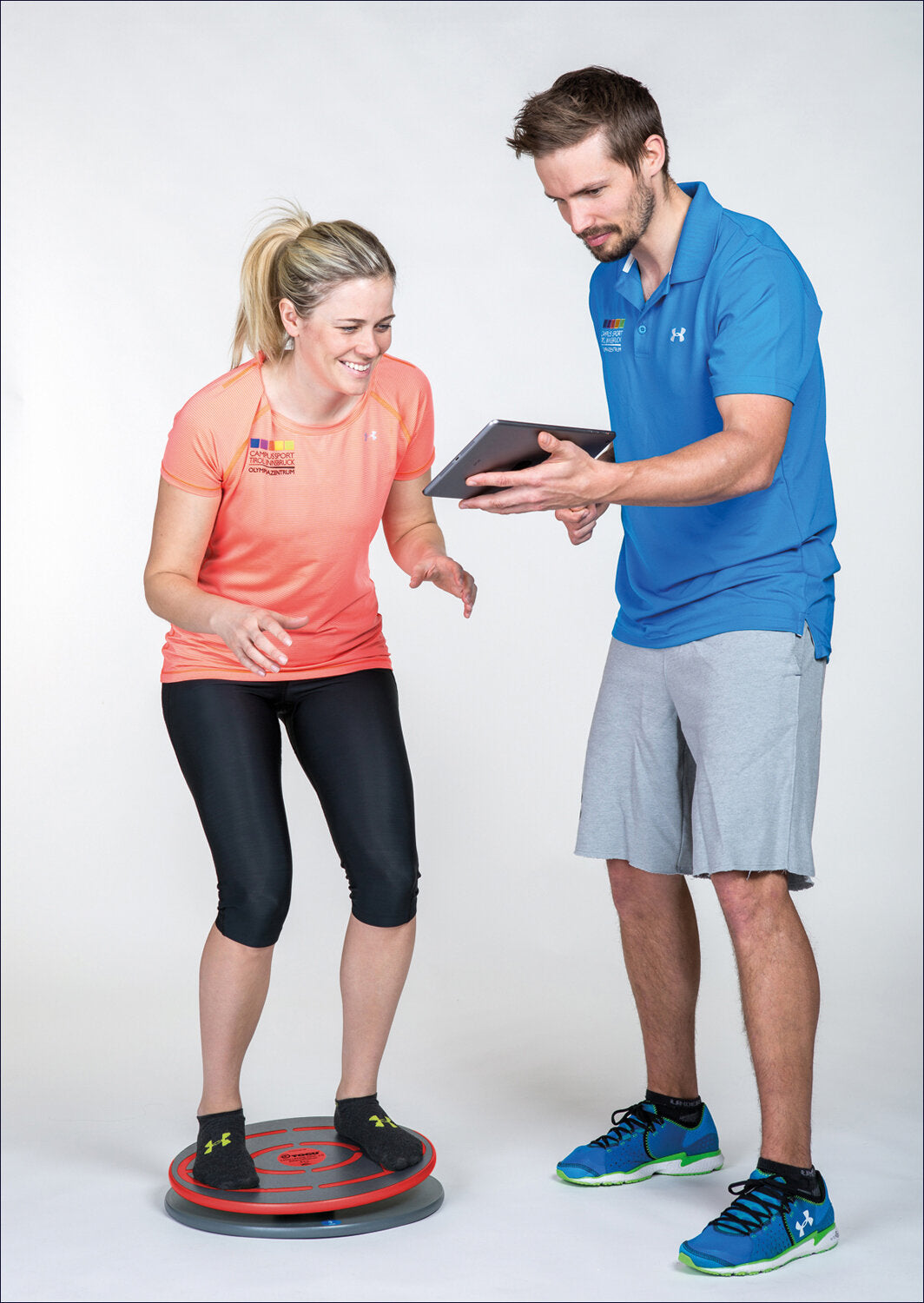 Frau steht auf einem Balance-Board, während ein Mann mit Tablet sie anleitet. Beide tragen Sportkleidung.