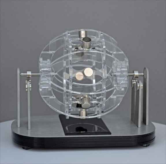 A transparent mechanical device with two white pills inside, mounted on a black base with metal components.