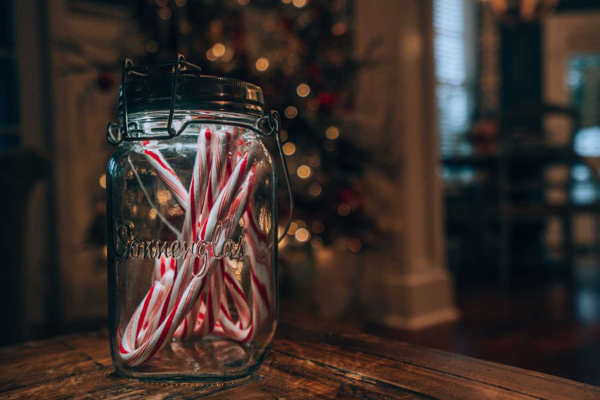 Glas mit Weihnachtslutschern auf einem Holztablett, im Hintergrund Weihnachtsbaum mit Lichtern.