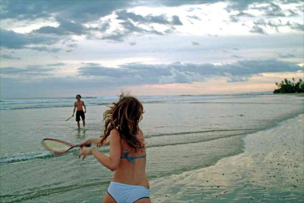 Ein Mädchen in Bikini spielt mit einem Tennisschläger am Strand, im Hintergrund ein Mann im Wasser, Wolken am Himmel.