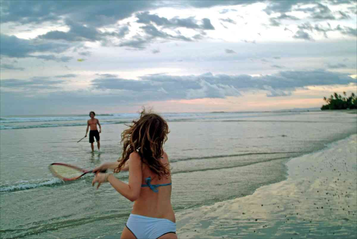 Ein Mädchen in Bikini spielt mit einem Tennisschläger am Strand, im Hintergrund ein Mann im Wasser, Wolken am Himmel.