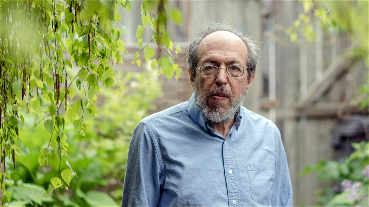 Älterer Mann mit Brille, grauem Bart und blauer Hemd im Garten, umgeben von grünen Pflanzen und Kletterpflanzen.