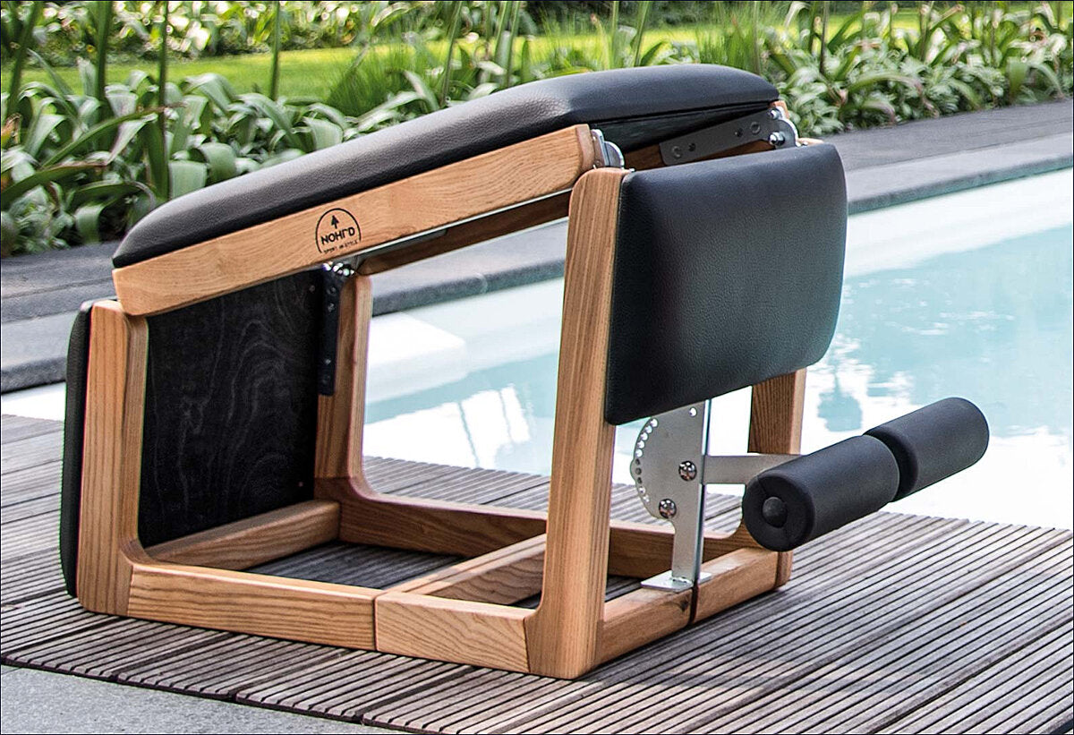 Fitnessgerät mit Holzrahmen, schwarzer Polsterung und Griff, neben Pool und Pflanzen.