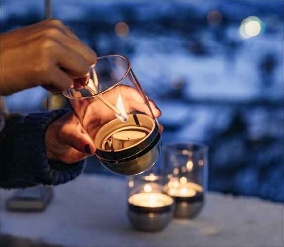 Hand zündet eine Kerze in einem Glas an, im Hintergrund sind weitere Kerzen auf einer Oberfläche.