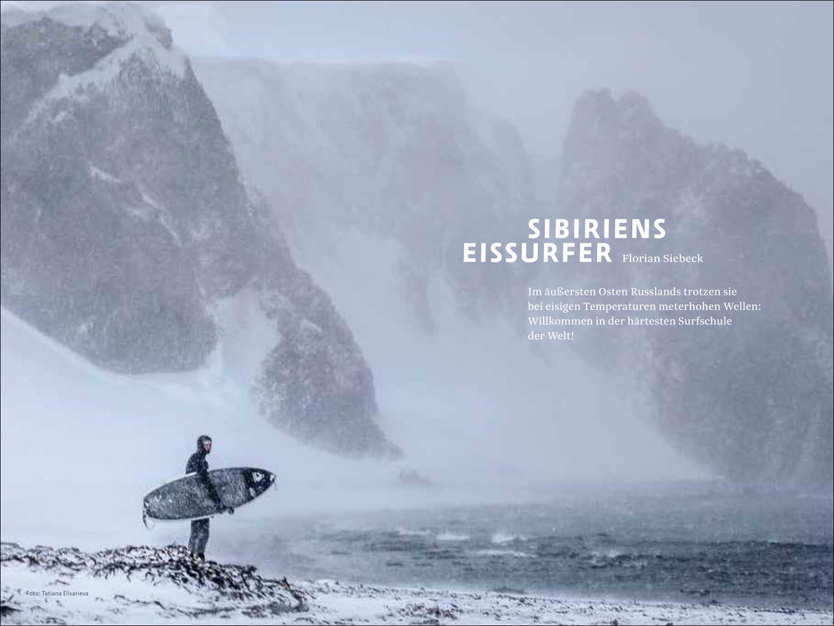 Ein einsamer Surfer mit Surfbrett steht im Schneesturm an einem eisigen Strand vor hohen, verschneiten Felsen.