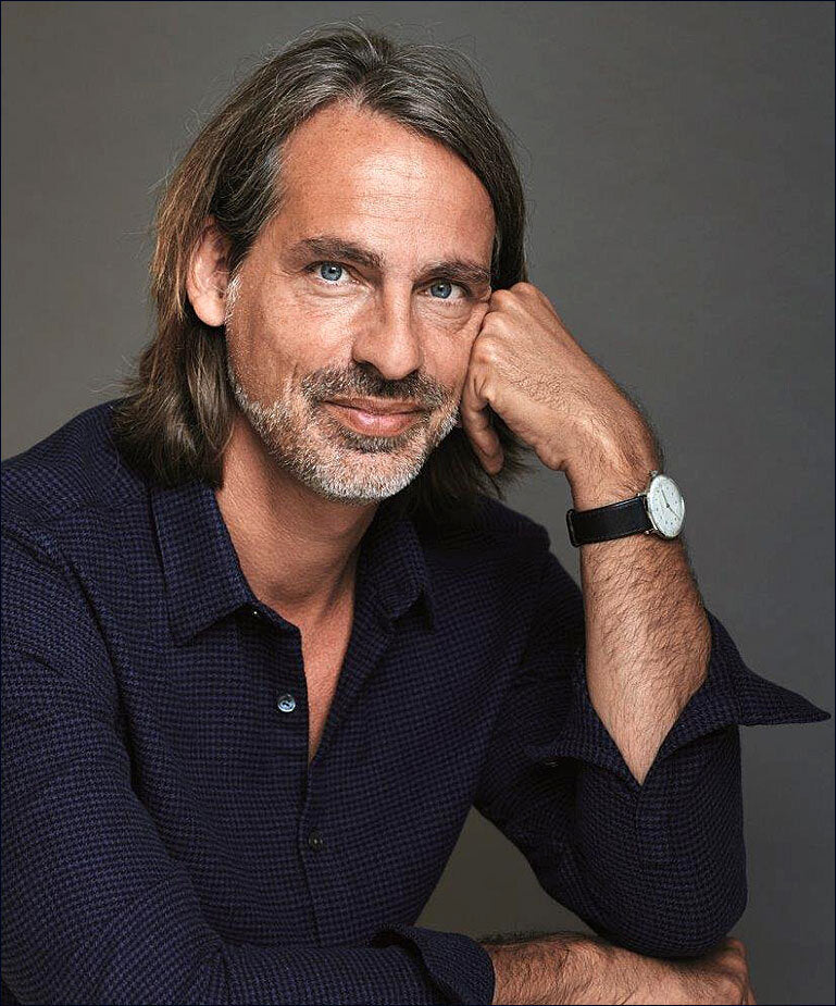 Porträt eines Mannes mit grauem Haar, Bart, blauen Augen, dunklem Hemd, Armbanduhr, vor grauem Hintergrund.