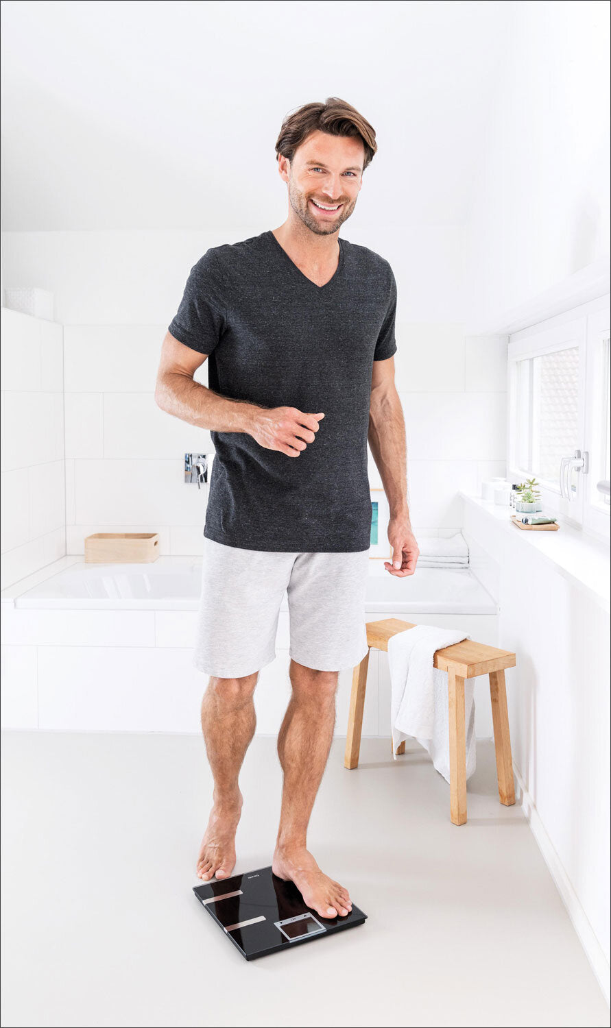 Mann steht auf einer Waage in einem weißen Badezimmer, trägt T-Shirt und Shorts, lächelt.