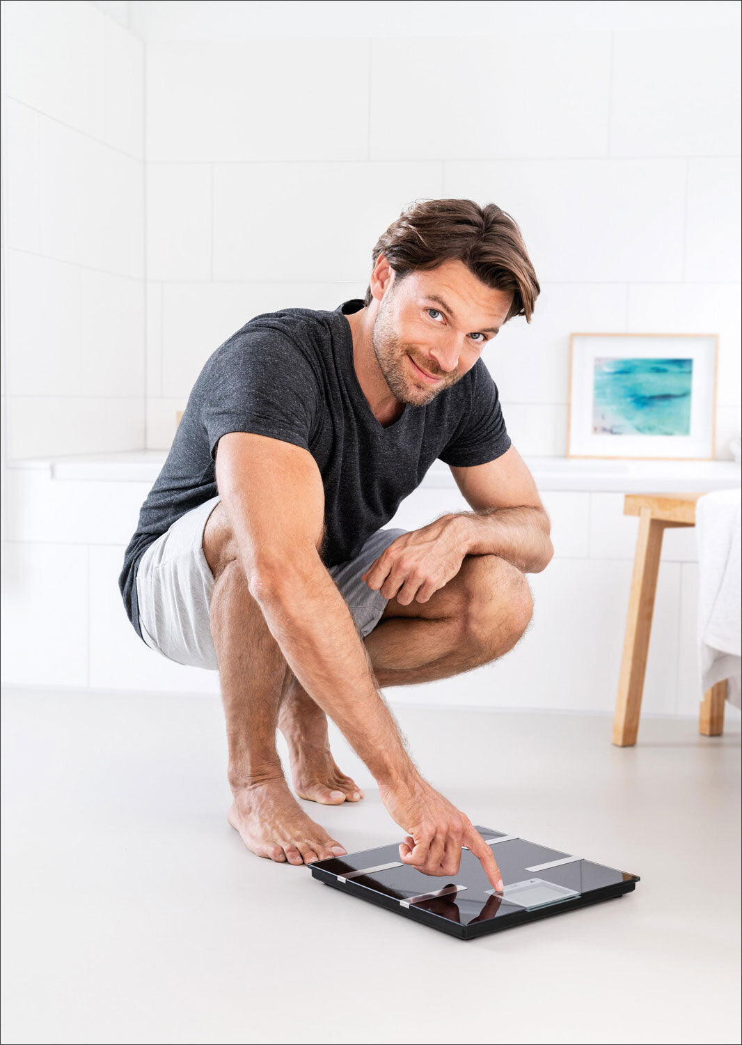 Mann steht auf Waage, zeigt auf Display, in weißem Raum mit Bild an Wand, sitzt auf Zehenspitzen, trägt T-Shirt und Shorts.