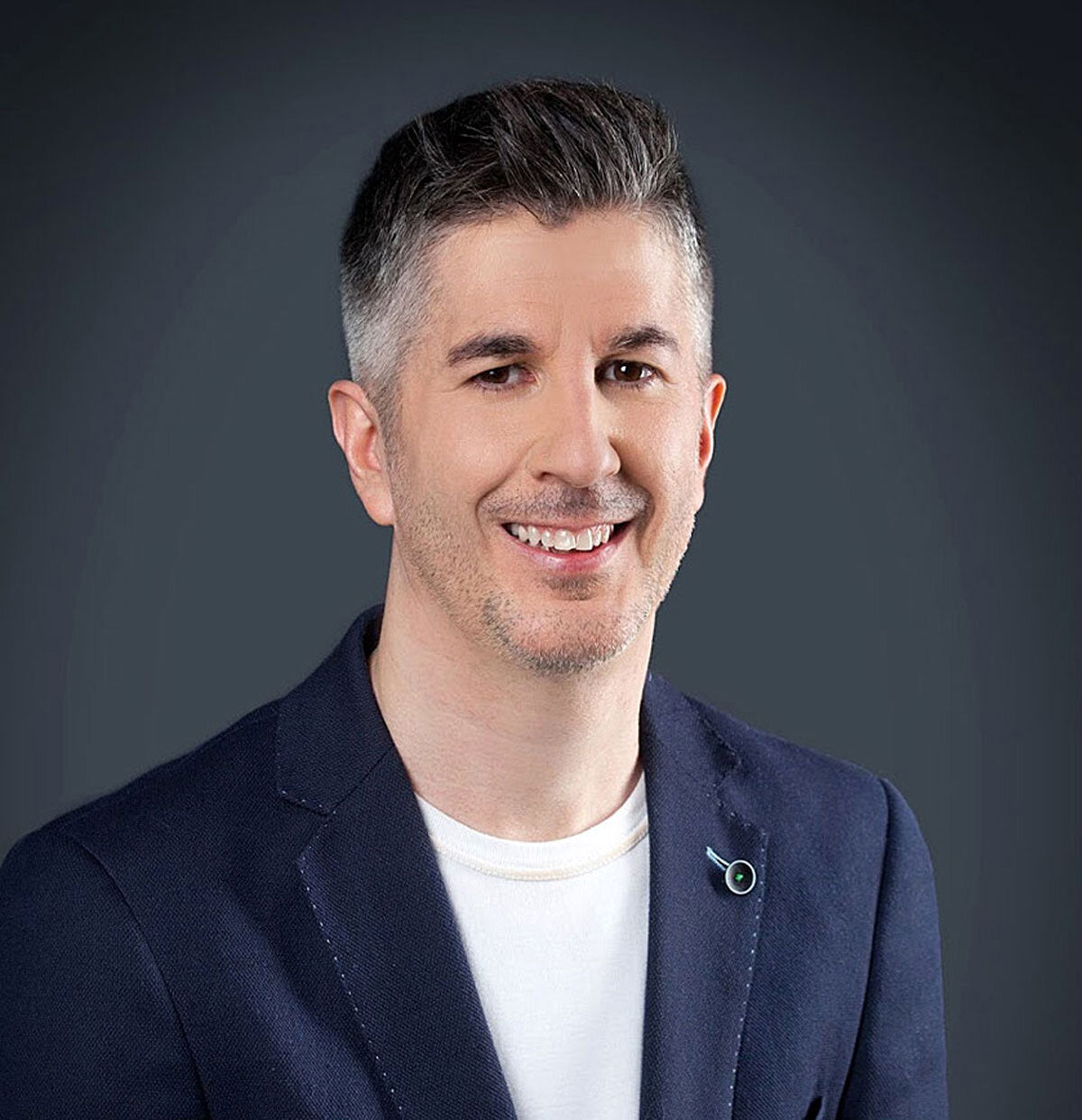 Mann mit dunklem Anzug, weißem T-Shirt, grauem Hintergrund, lächelnd, kurze, graue Haare.