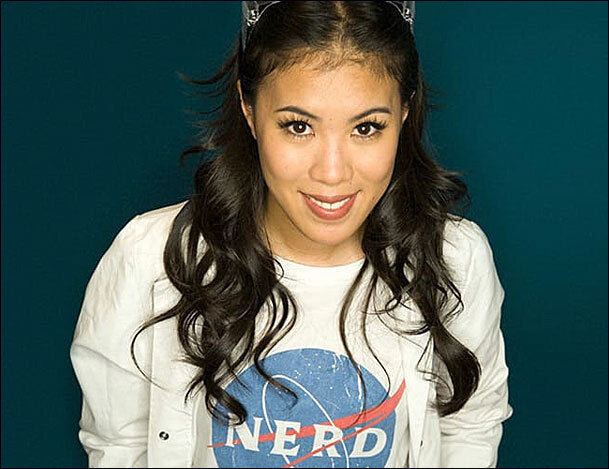 Junge Frau mit langen, dunklen Haaren, trägt ein weißes T-Shirt mit NASA-Logo, lächelt, dunkler Hintergrund.