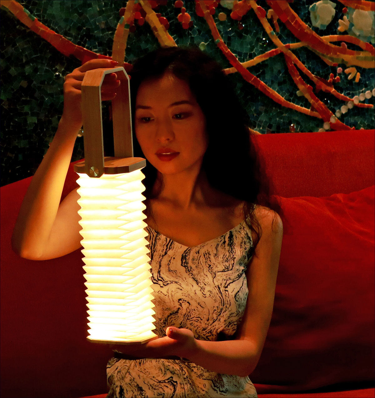 Frau hält eine leuchtende, spiralförmige Lampe vor sich, sitzt auf rotem Sofa vor einer bunten, mosaikartigen Wand.