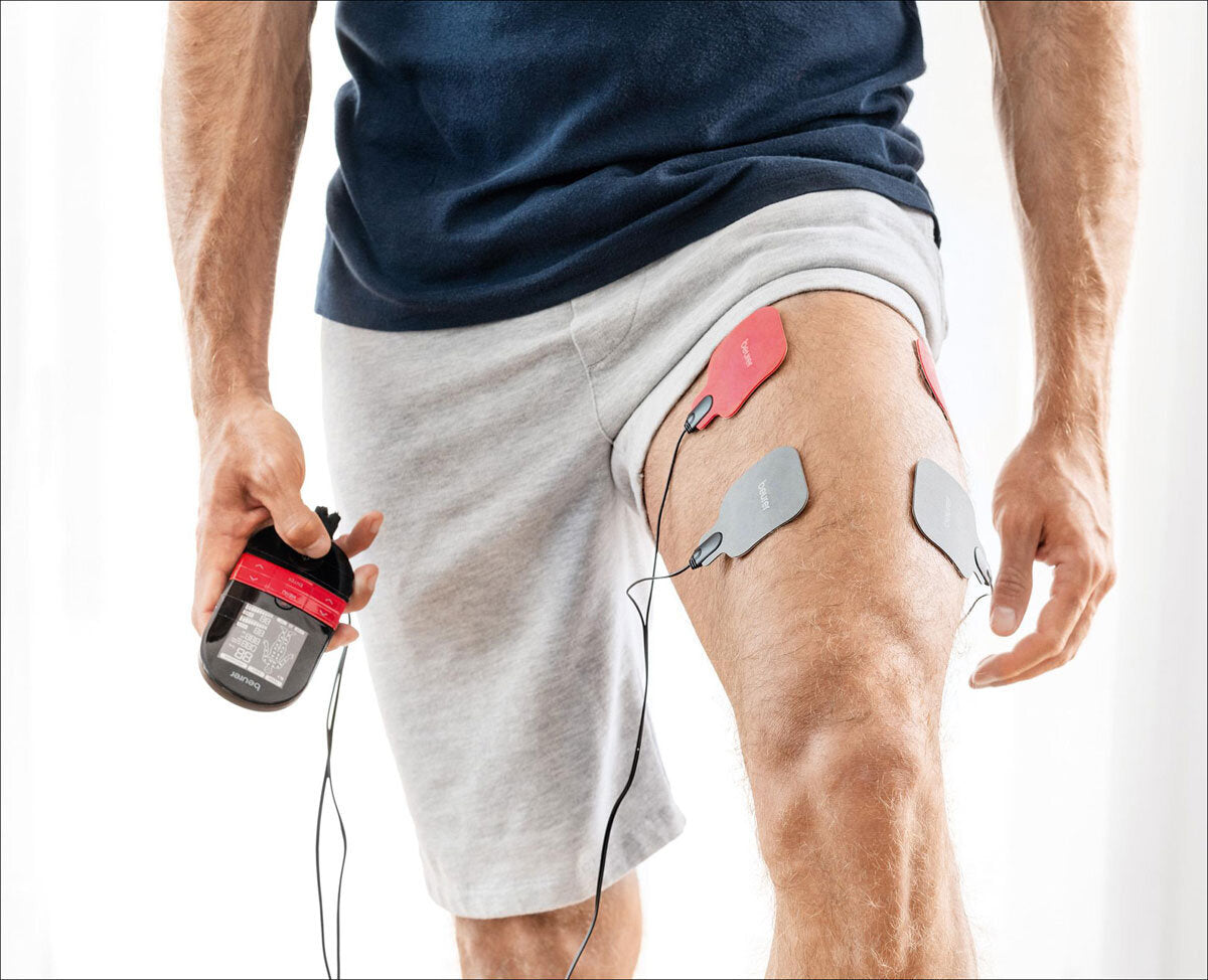 Mann mit EKG-Elektroden an Oberschenkel, hält EKG-Gerät in Hand, trägt graue Shorts und blauen T-Shirt.