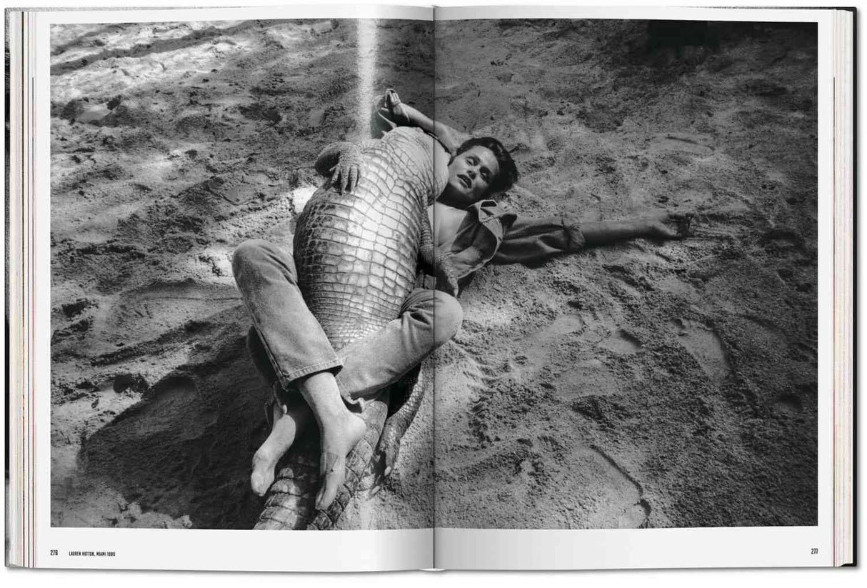 Frau liegt mit einem Krokodil auf sandigem Boden, umarmt es, Blick zur Kamera, in Schwarz-Weiß.