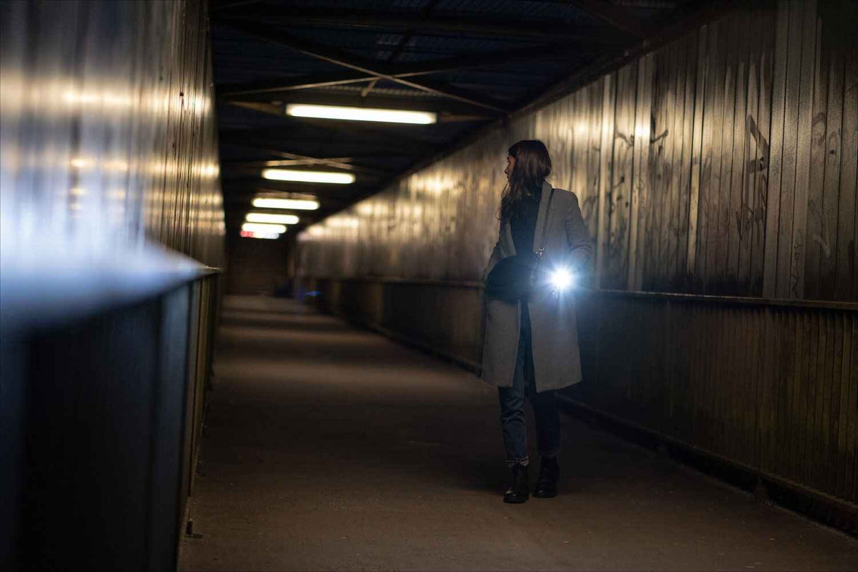 Frau steht in einem dunklen Tunnel, hält eine Tasche, trägt einen Mantel, und hält eine Taschenlampe.