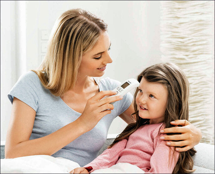 Frau hält ein Fieberthermometer an die Stirn eines lachenden Mädchens.