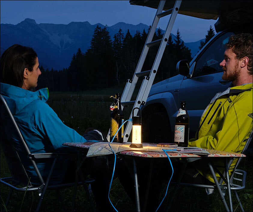 Zwei Personen sitzen abends bei einem Camping-Tisch mit Lampen, Bierflaschen, vor einem Van, im Hintergrund Berge und Bäume.
