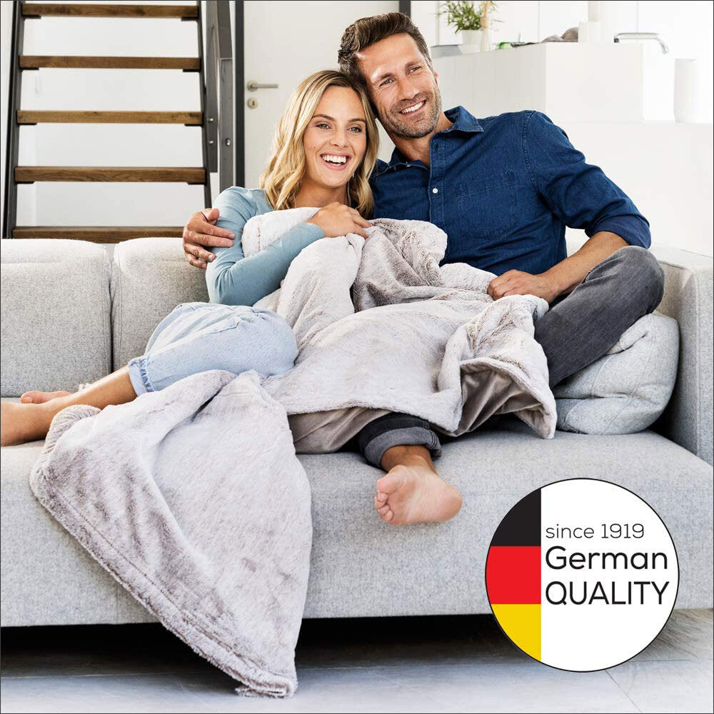 Ein lachendes Paar sitzt auf einem Sofa, umgeben von Decken, im hellen Wohnzimmer. Im unteren rechten Bildrand ein Logo