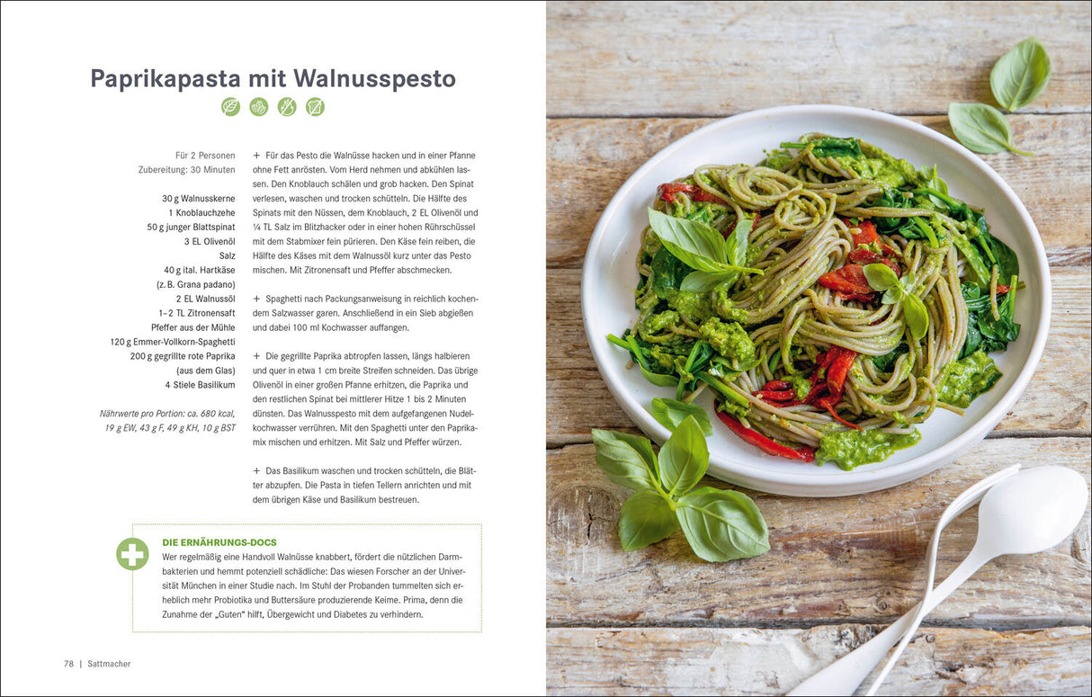 Bunter Pasta-Teller mit grünen, roten und braunen Zutaten auf einem Holztisch, daneben ein weißer Löffel.