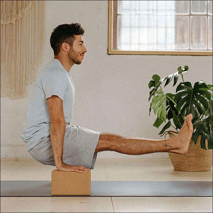 Ein Mann sitzt auf einem Yoga-Block, streckt ein Bein auf eine Pflanze, im Hintergrund ein Fenster.