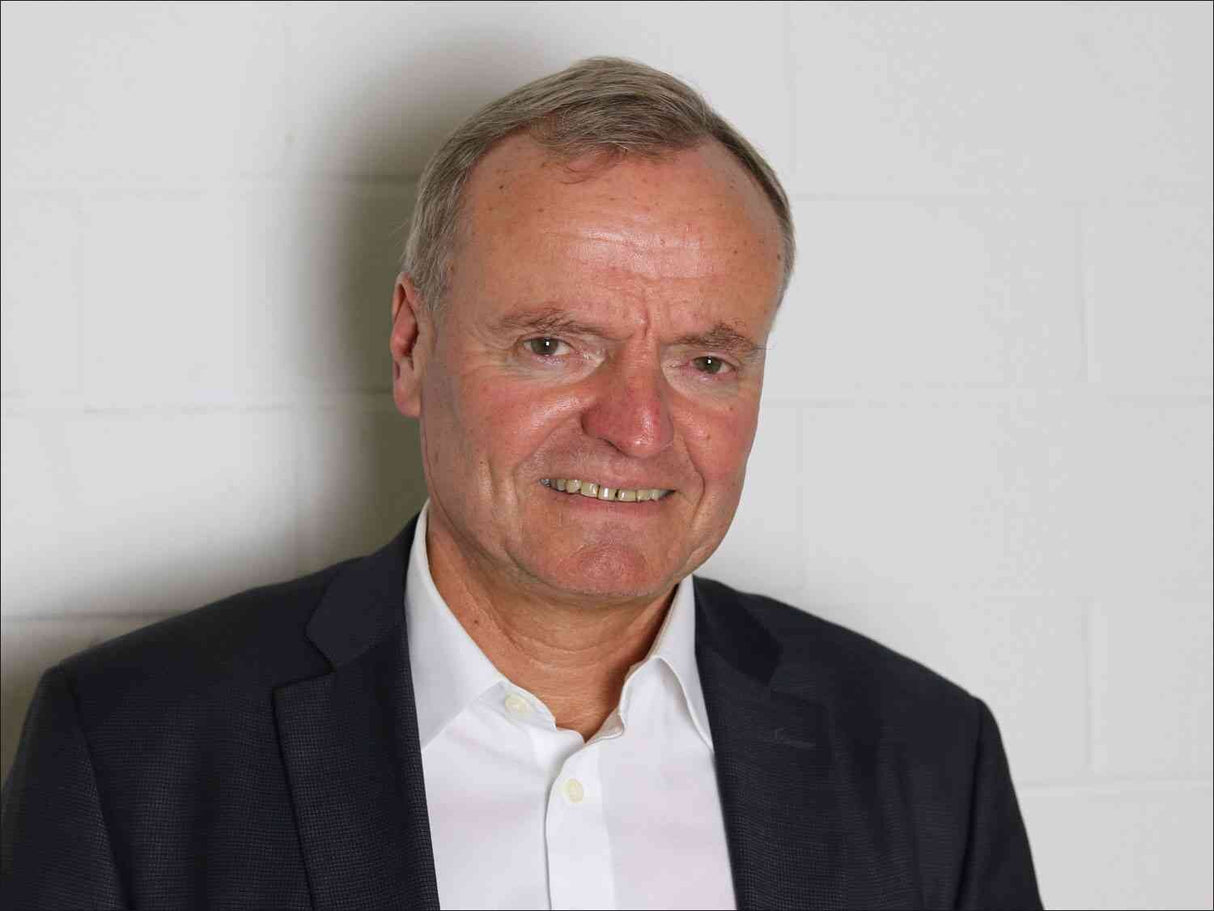 Porträt eines älteren Mannes in Anzug und weißem Hemd vor weißer Wand.