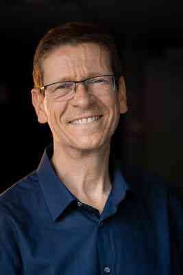 Mann mit Brille, lächelnd, trägt dunkles Hemd, dunkler Hintergrund.