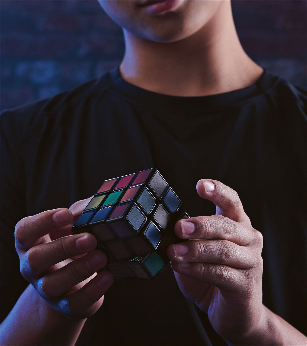 Person hält einen aufgemalten Rubik's Cube in den Händen, trägt ein schwarzes T-Shirt, dunkler Hintergrund.
