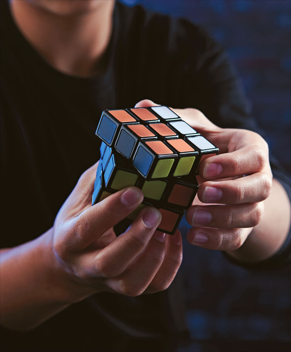 Person hält einen aufgerissenen Rubik's Cube in den Händen, dunkler Hintergrund, schwarze Kleidung.